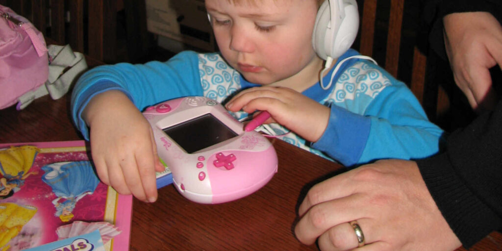 A small boy wearing headphones with a pink Leapster. A parent watches on. 