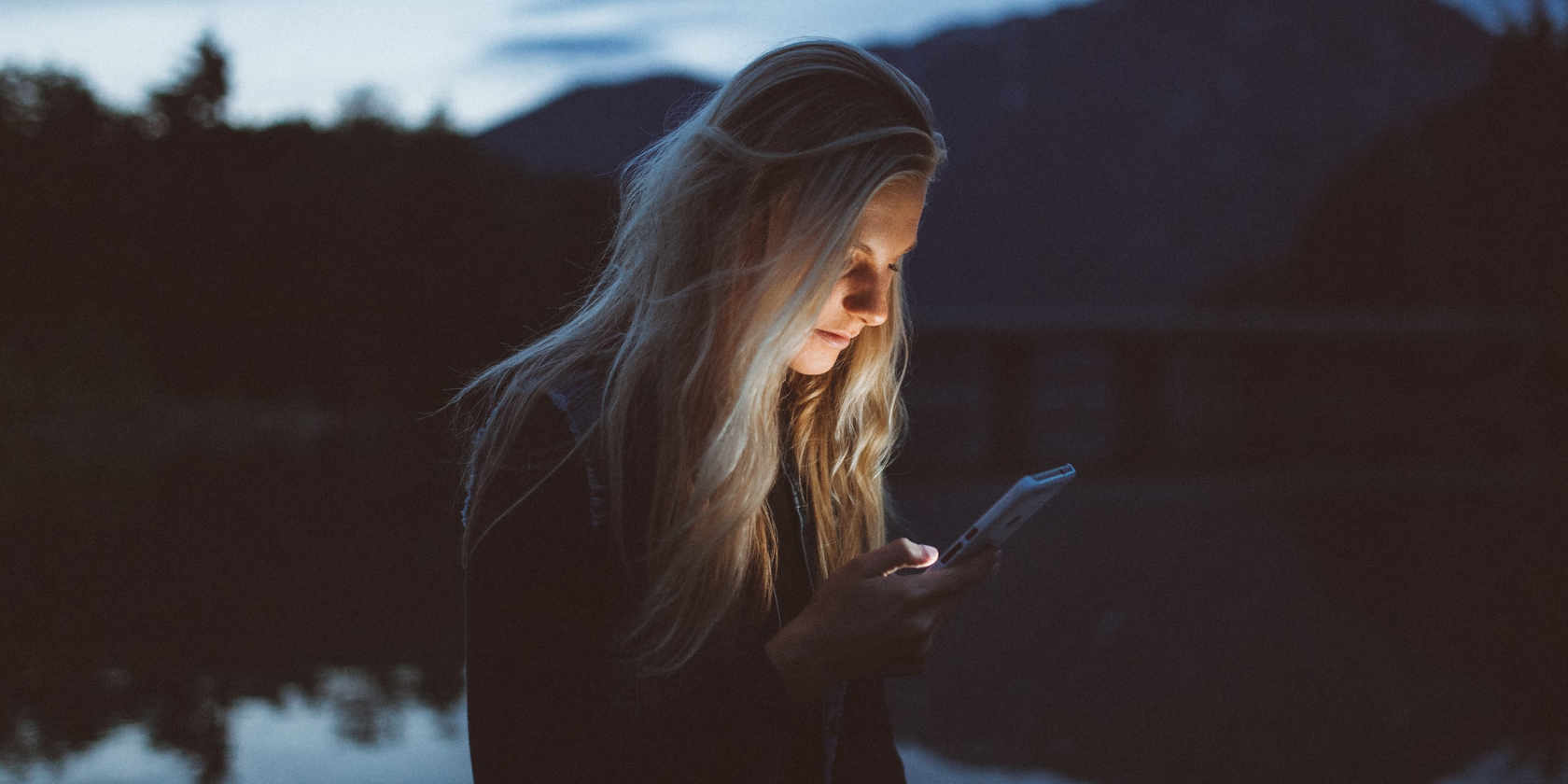 A blonde woman at night by a lake. Her face is illuminated by the light from her phone
