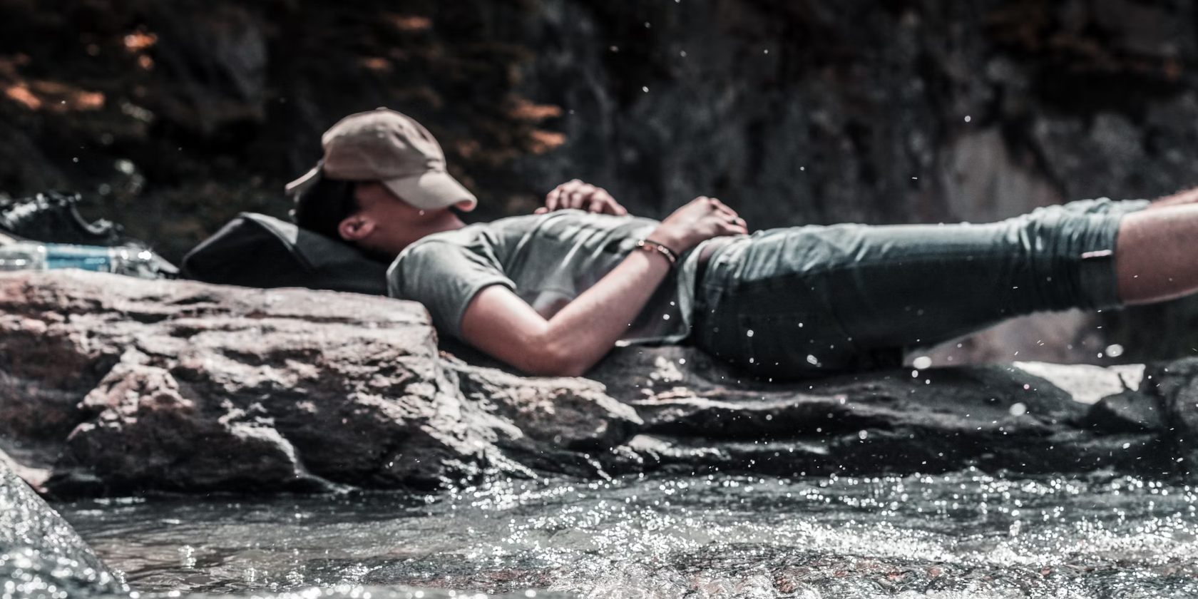 A man asleep on a rock in the middle of a river. His cap is over his face