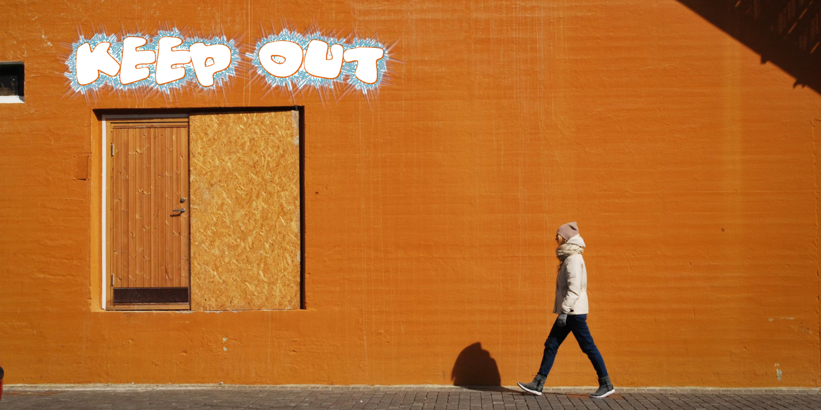 A person walking past an orange wall with an accessible door a few weeks above ground. The words Keep Out are sprayed above the door