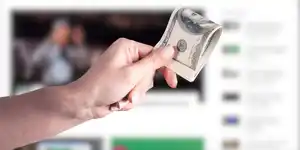 A hand holding folded money in front of a blurred web page