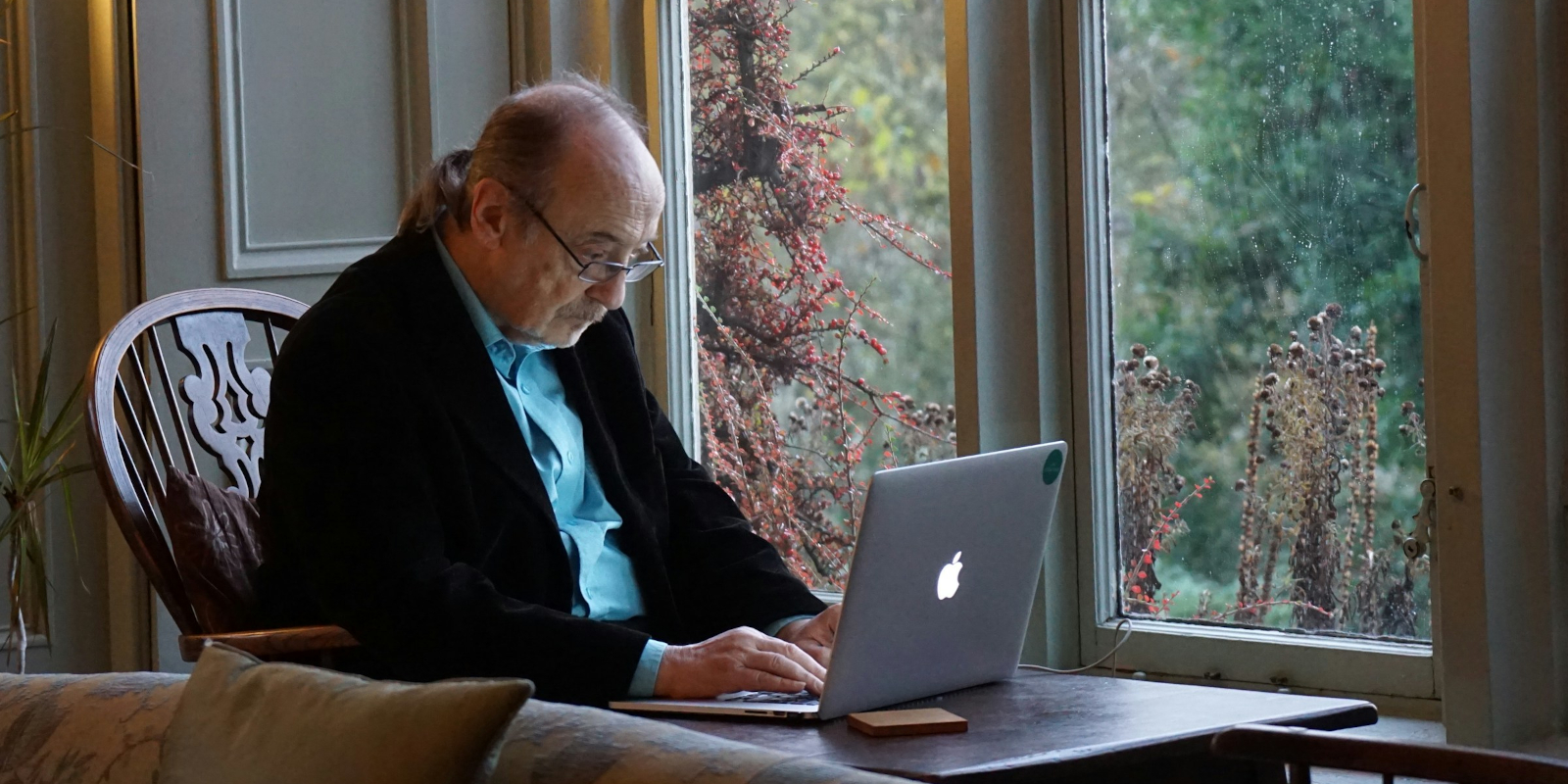 An older man with a ponytail using a laptop in front of a window